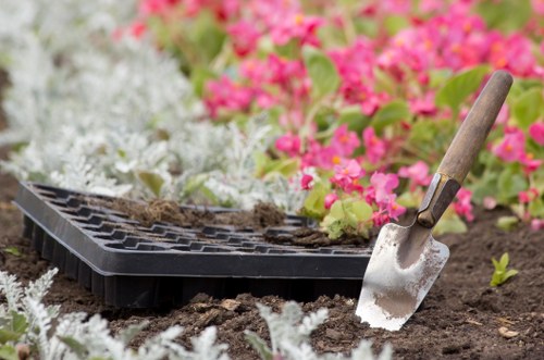 The image depicts a gardening scene in a well-maintained backyard, with a close-up of a small trowel inserted into dark, moist soil, suggesting recent planting or soil work. In the background, there are vibrant, densely planted flower beds with pink and white flowers, contrasting against lush green foliage. The flower beds are well-defined, edged with a neat border, and the garden area shows a healthy, organized landscape typical of a professional gardening service. A black plastic seed tray with soil remnants rests on the ground nearby. The setting appears to be outdoors on a bright and clear day, with natural lighting highlighting the rich colours of the flowers and the soil's texture. The overall scene exemplifies typical garden maintenance and planting activities that a landscaping or garden care provider such as Hedge Trimming Sydenham might perform within a suburban garden in the SE postcode area, reinforcing their landscaping and gardening expertise.