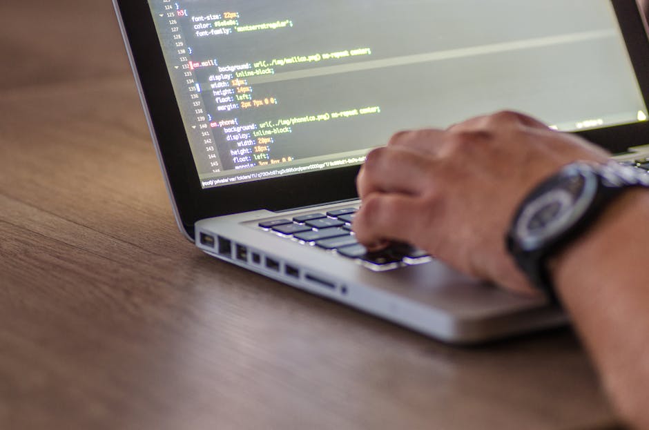 A close-up view of a person's hand, wearing a black wristwatch, typing on a silver laptop placed on a wooden desk. The laptop screen displays lines of code with syntax highlighting; the background is