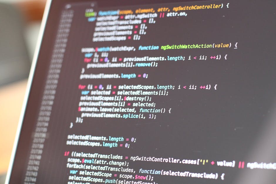 A close-up view of a person's hand, wearing a black wristwatch, typing on a silver laptop placed on a wooden desk. The laptop screen displays lines of code with syntax highlighting; the background is